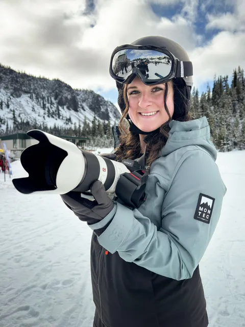 Marin on a snowy mountain in ski gear, holding a telephoto lens